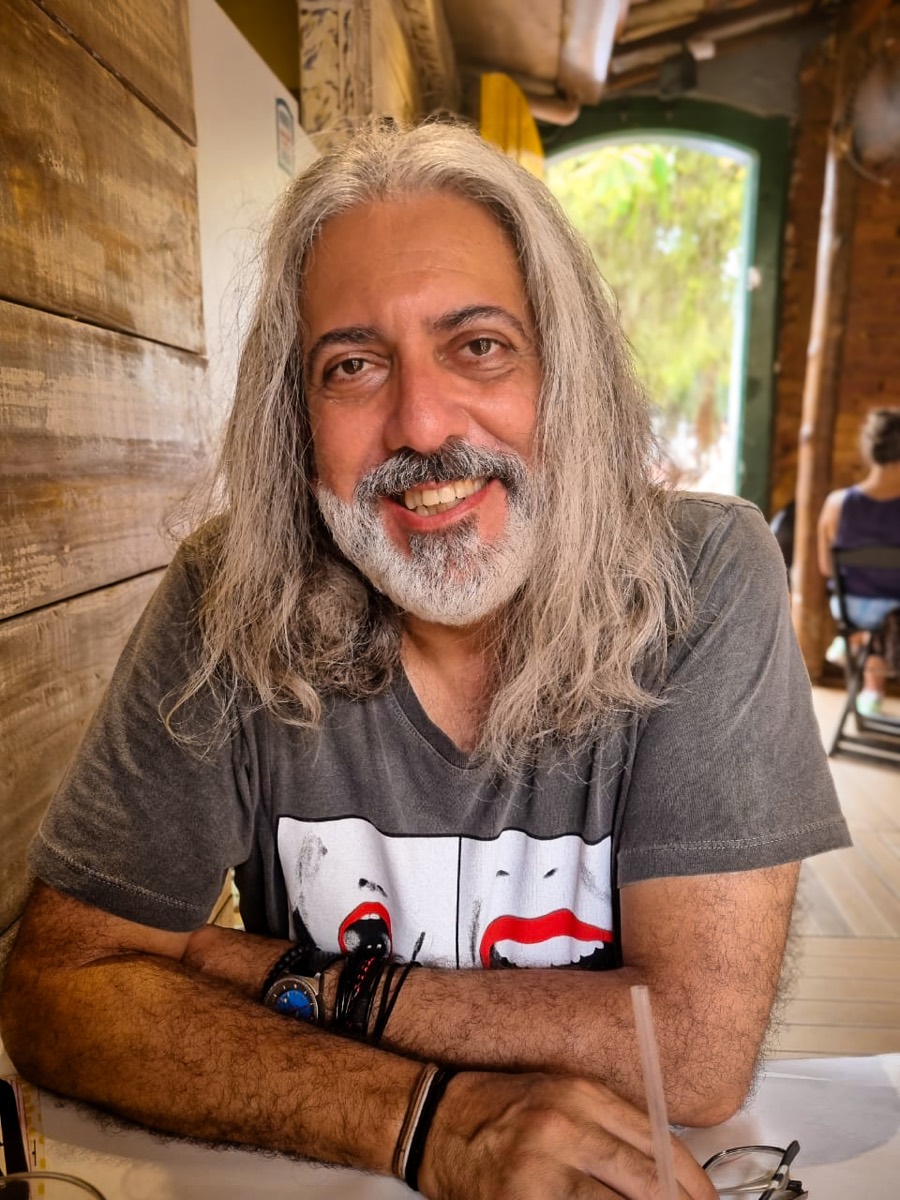 Montalvo Machado, artista brasileiro de cabelos grisalhos longos, sorrindo, sentado à mesa.
